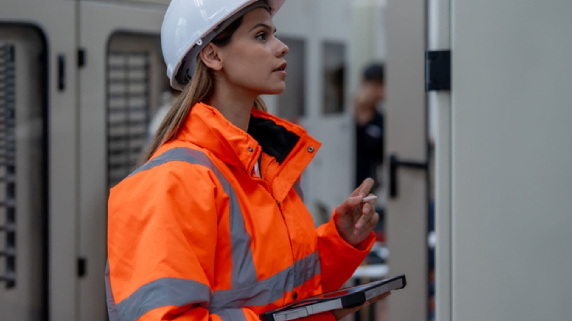 Female technician in safety gear inspects control panel with tablet in hand at an industrial facility for maintenance check.