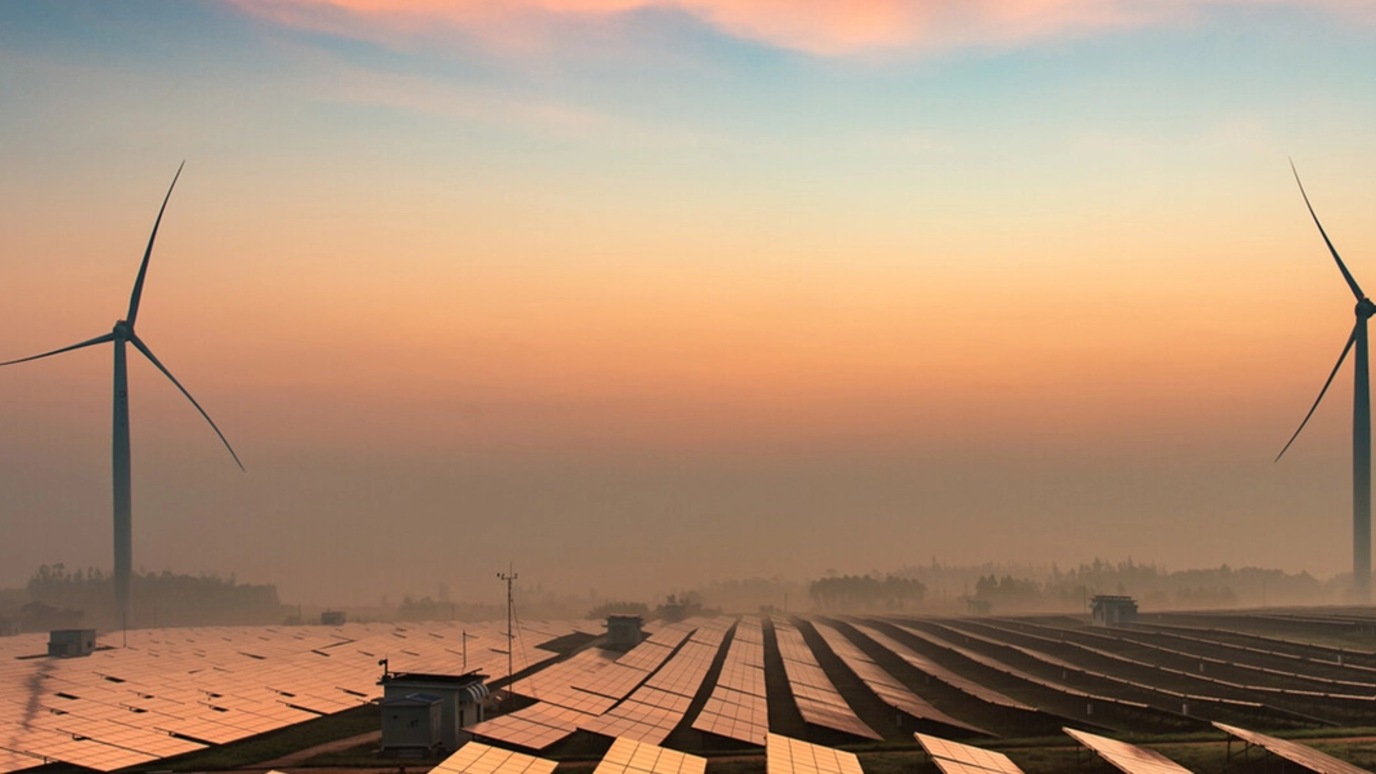 A picture of two wind turbines and many solar panels in a morning haze.