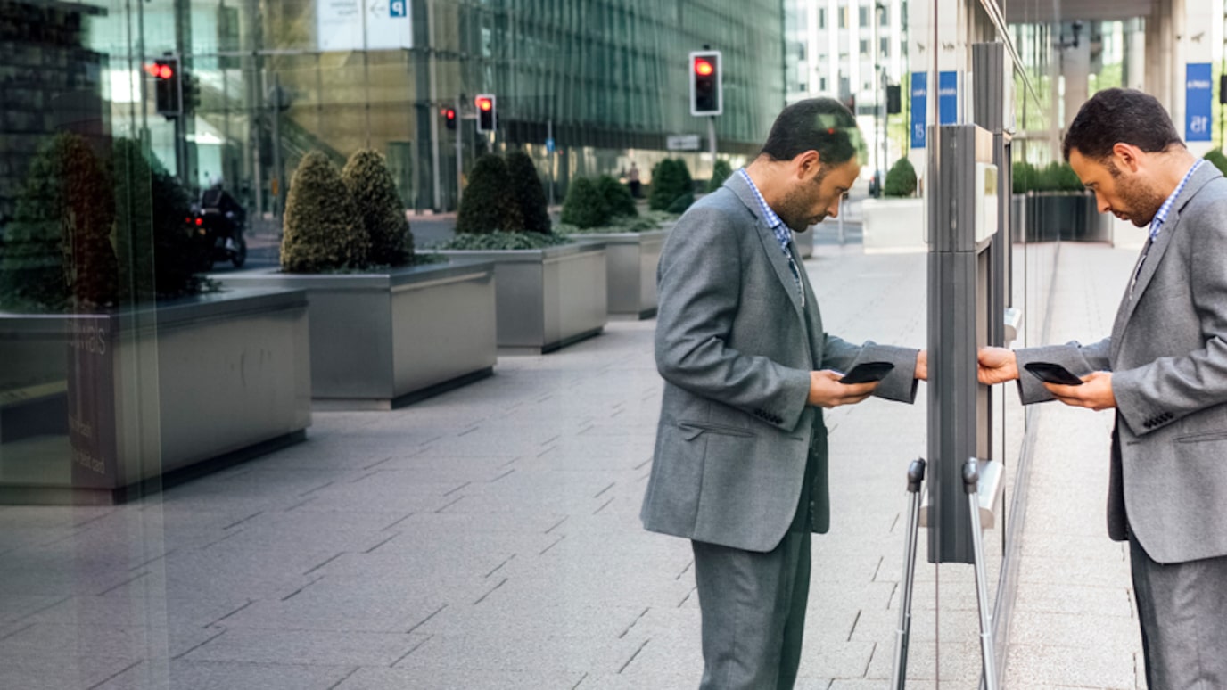 A person standing at an ATM with their reflection showing on a glass wall next to the ATM.