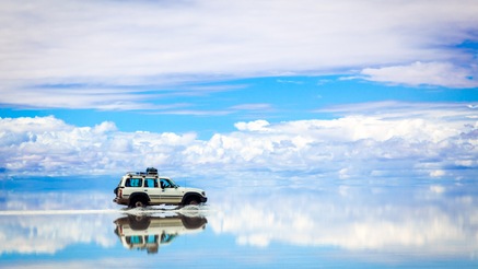 A car driving in shallow water with clouds reflecting on the surface of the water.