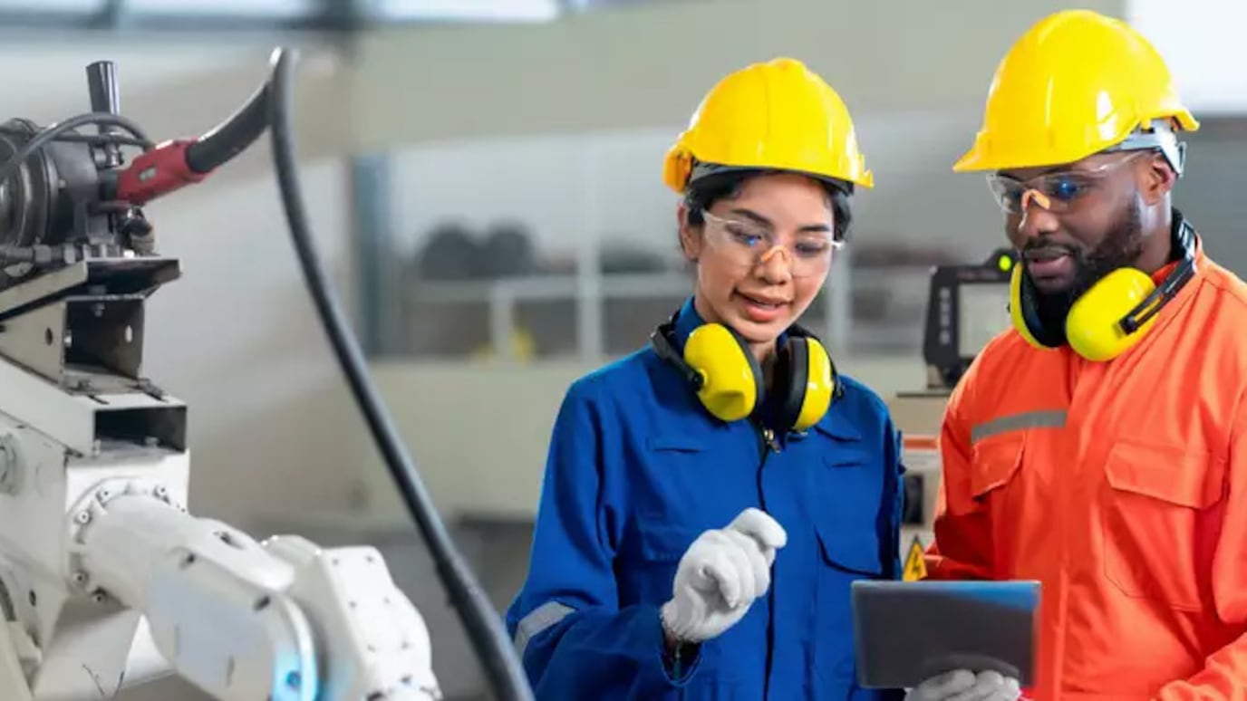 Two engineers in overalls and hard hats look at tablet next to robotic arm