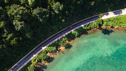 An aerial view of a winding road along a coastline, with lush green trees on one side and clear turquoise water on the other. A single car is visible driving along the road.