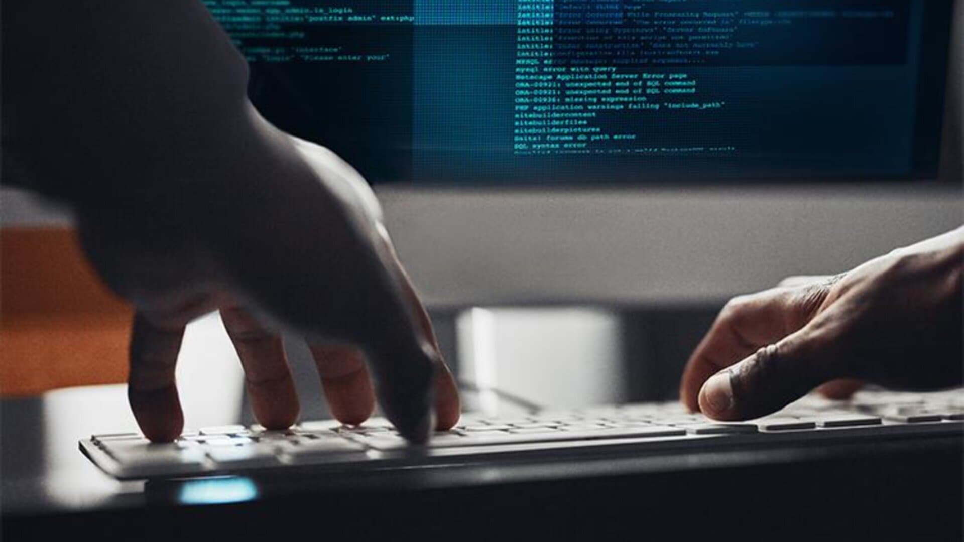 Close-up of hands typing on a keyboard with a computer screen displaying lines of code in the background