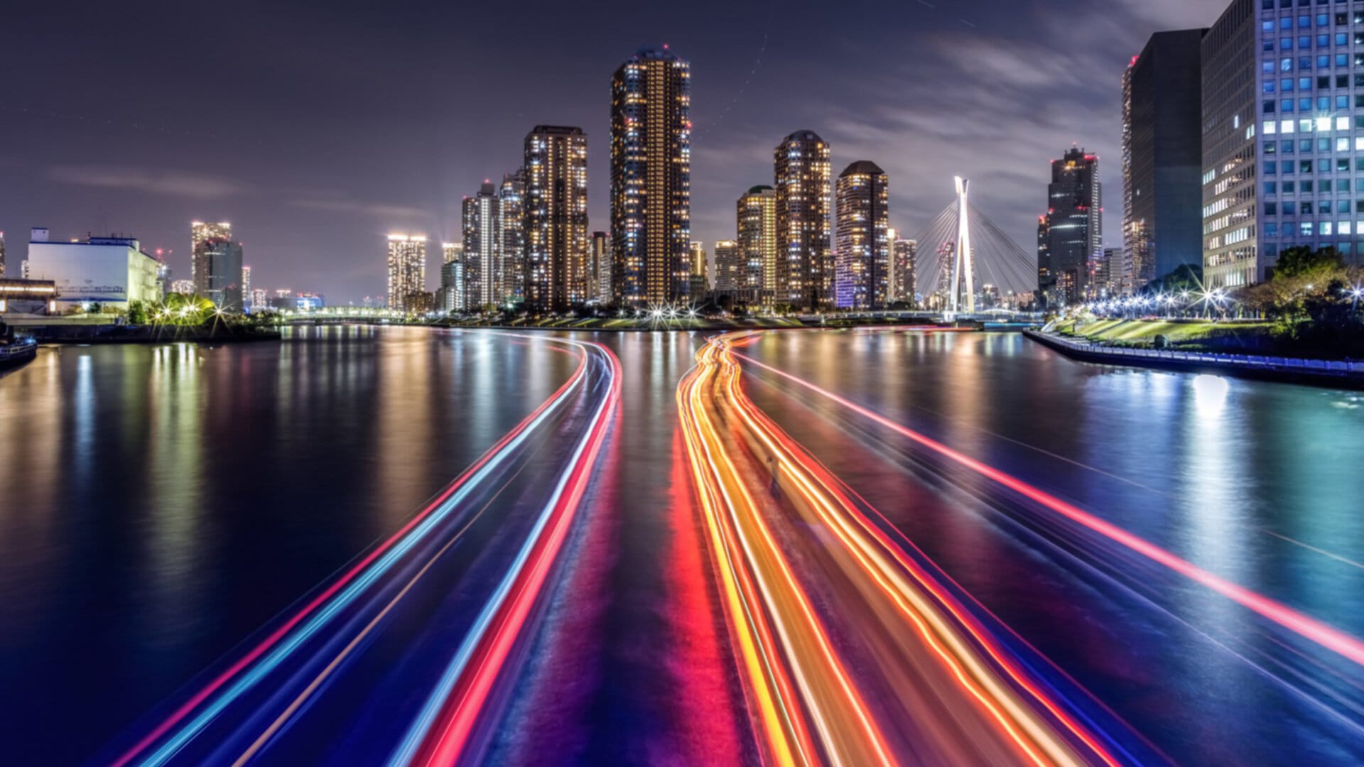 A night time view of a city skyline with a river in the foreground. The river is illuminated by streaks of light from boats passing by. The city skyline is lit up with lights from buildings and streetlights