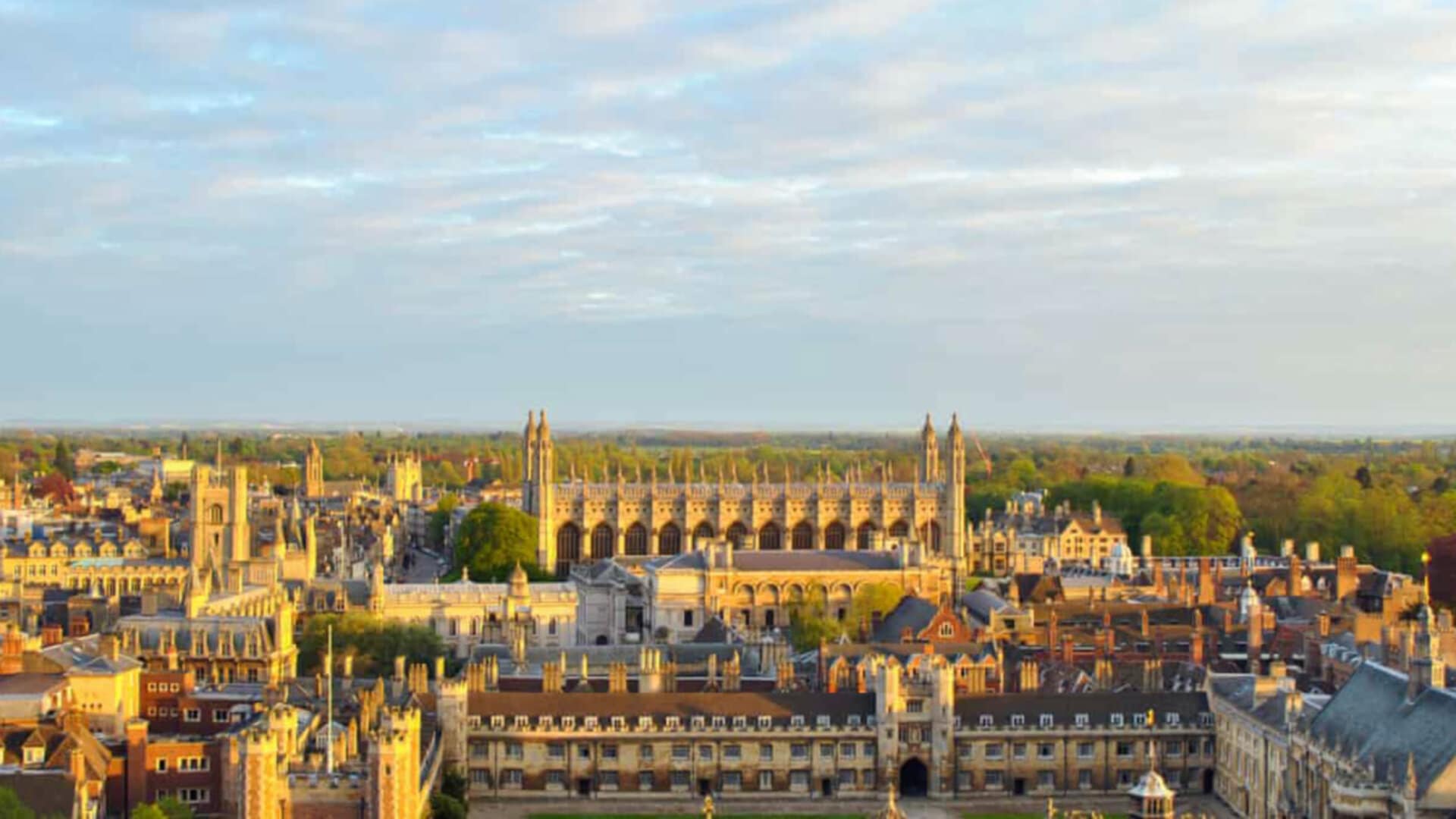 A cityscape of Cambridge, featuring historic buildings and lush greenery under a partly cloudy sky