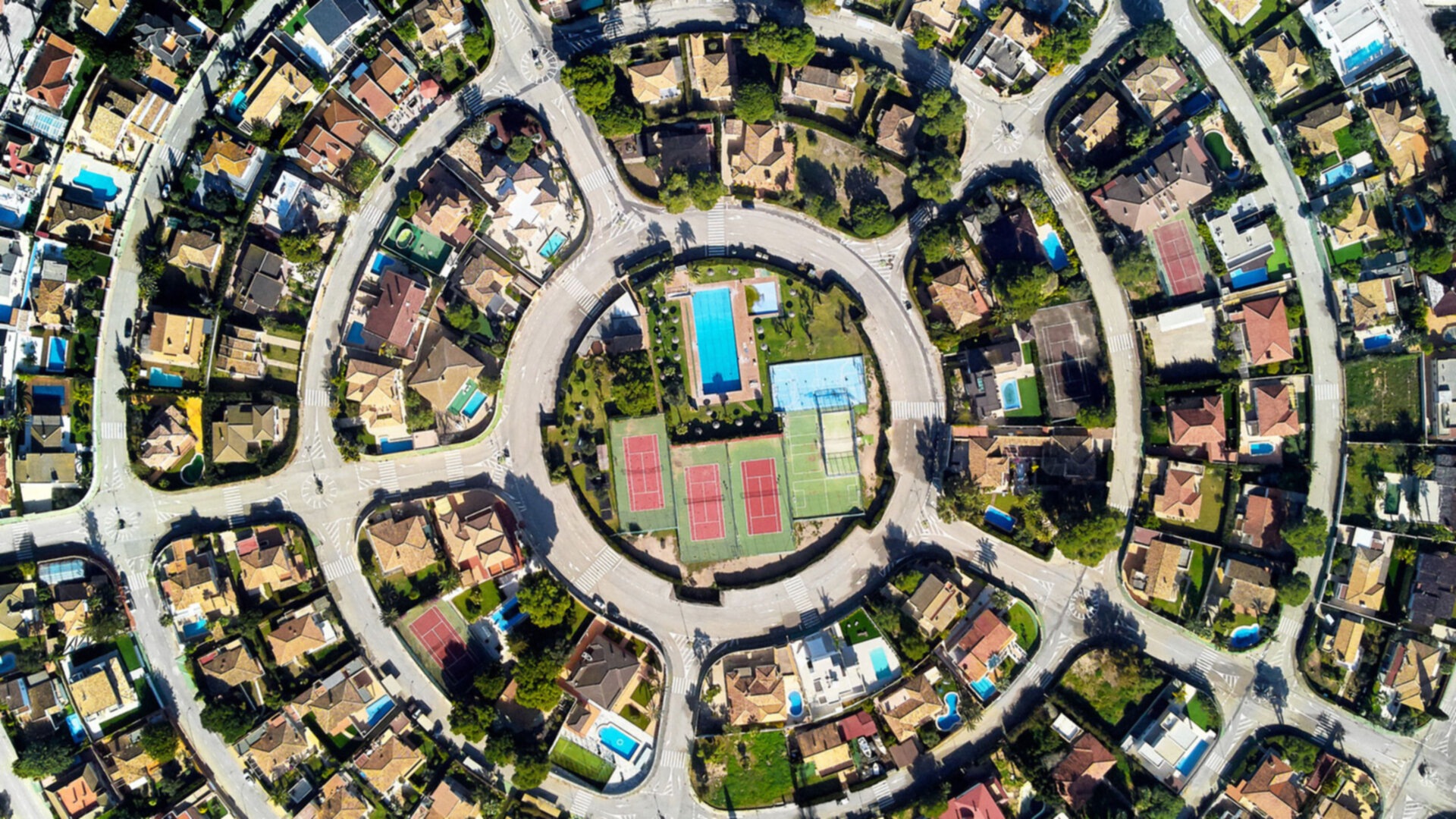 An aerial view of a suburban neighborhood with a circular road surrounding a park with tennis courts and swimming pools