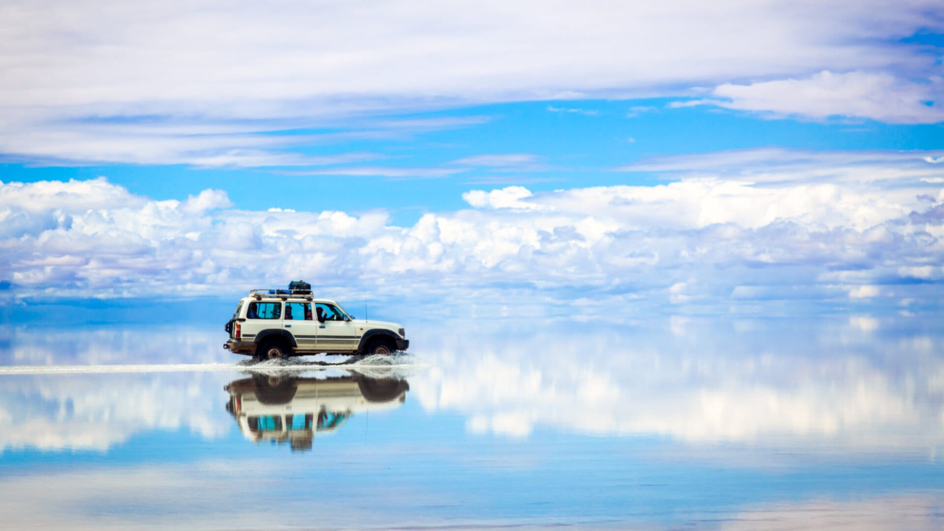 A stunning landscape of the Salar de Uyuni salt flats in Bolivia. A white SUV is driving through shallow water, creating a perfect reflection on the mirror-like surface.