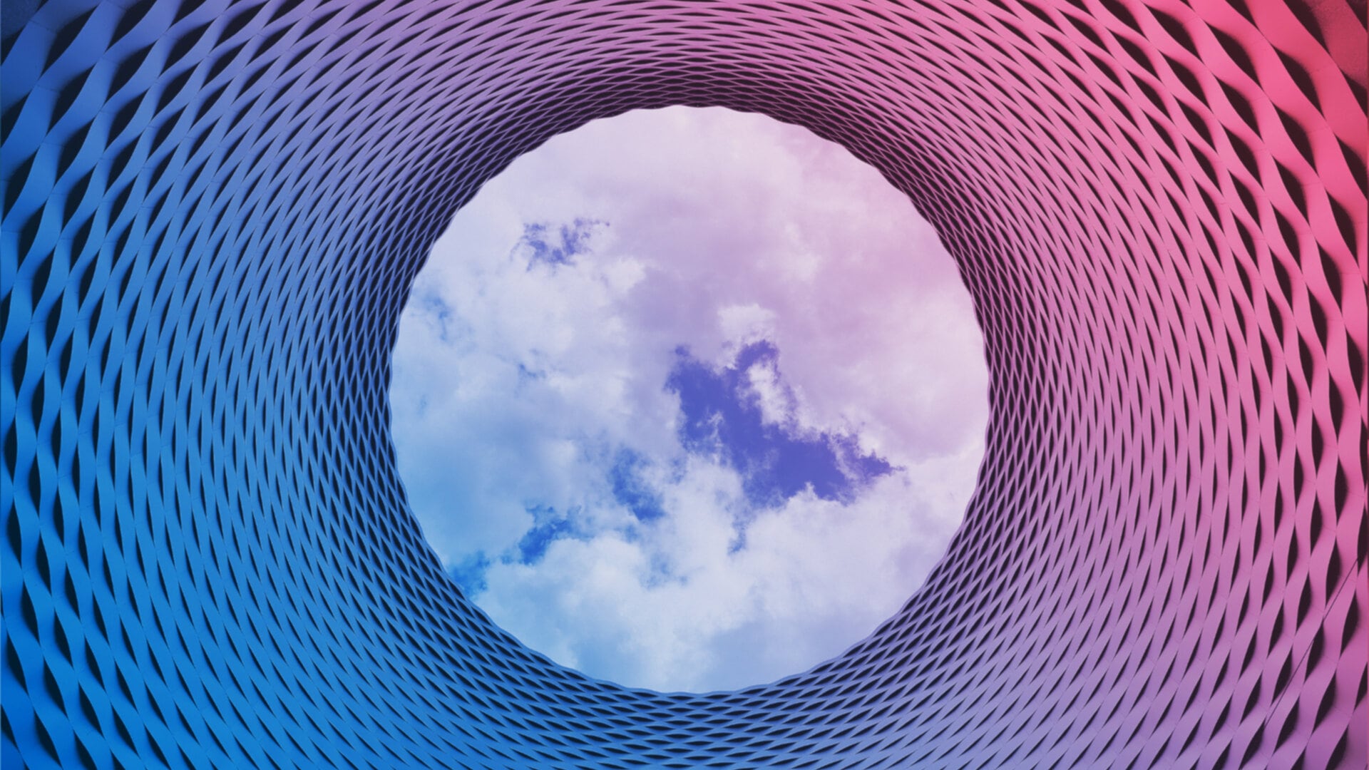 A close-up view of a curved, patterned building facade with a circular opening revealing a blue sky with white clouds