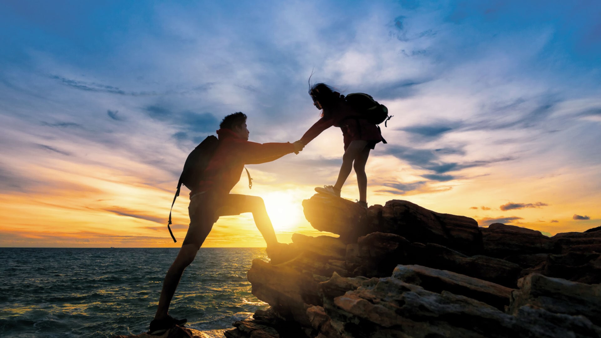 Two silhouetted figures engaged in a hiking or climbing activity on a rocky coastal area during a dramatic sunset. One person is reaching up to help the other climb onto a higher rock formation.