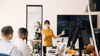 A woman presenting to a group of colleagues in a modern office setting. She is pointing at a large screen displaying code.