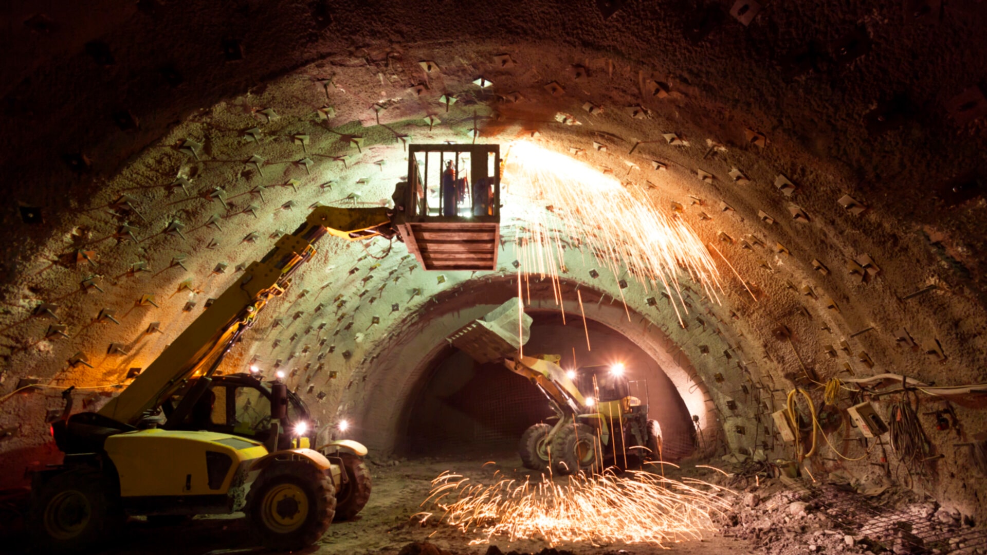 An underground construction: Arched tunnel with workers on suspended platform welding, sparks flying.