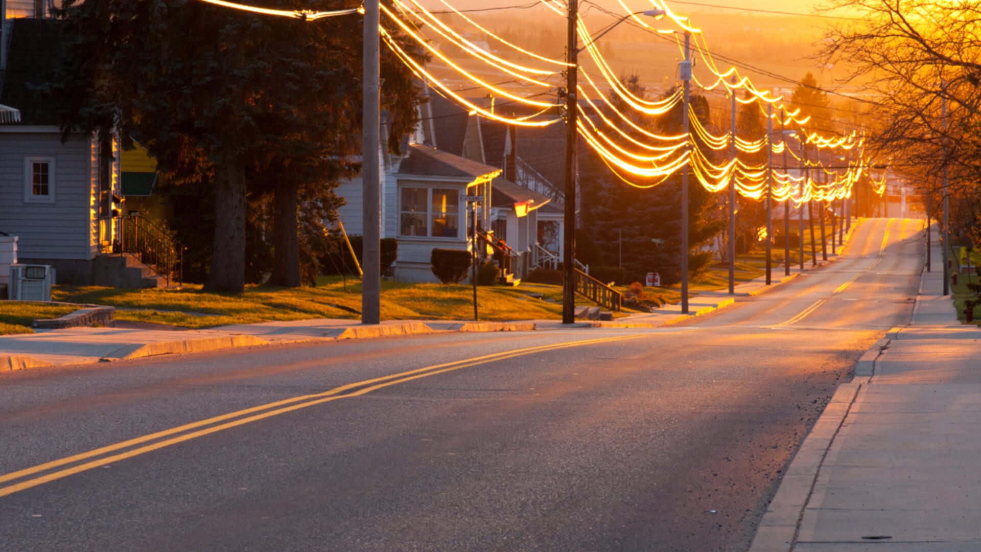 A quiet residential street at sunset. The street is lined with houses and utility poles, with power lines glowing warmly in the evening light.