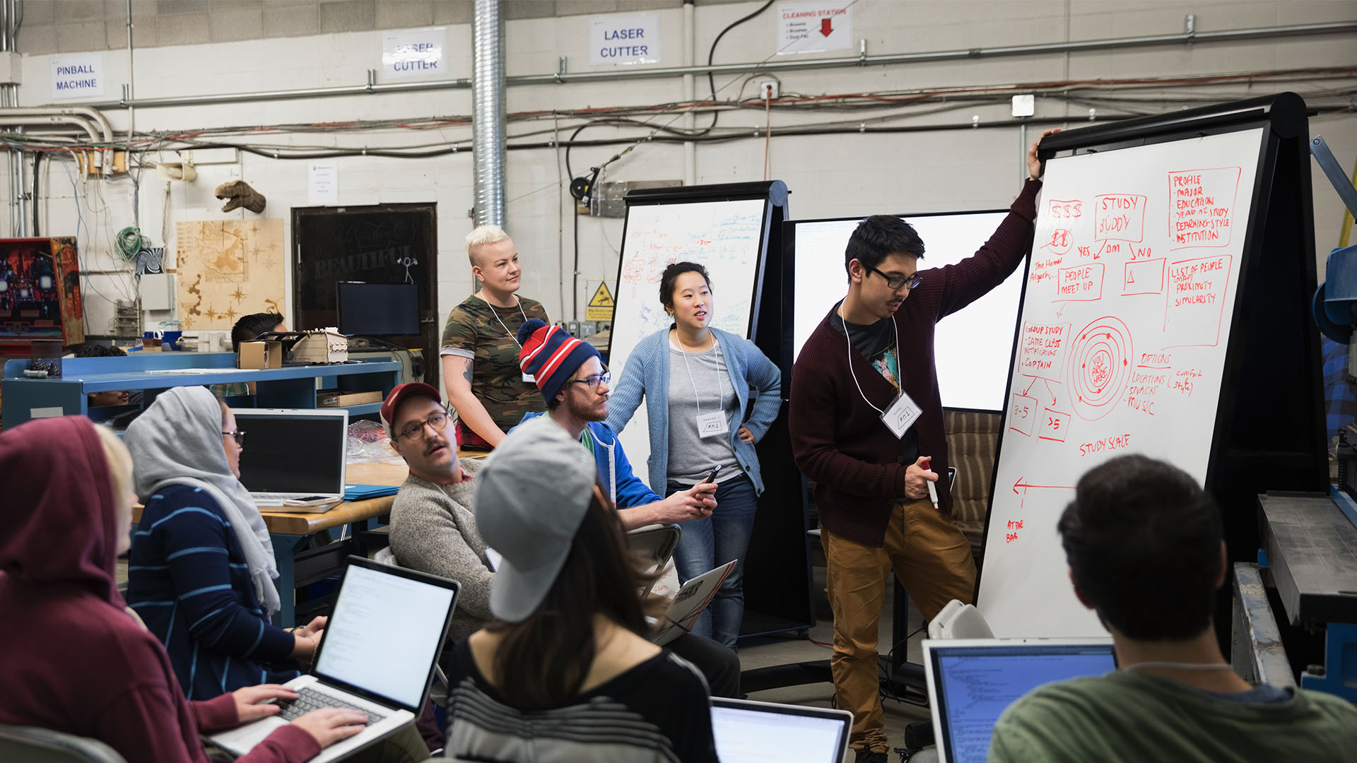 A group of people gathered around a whiteboard in a workshop setting. One person is presenting a diagram about a study buddy system, while others listen and take notes on their laptops.