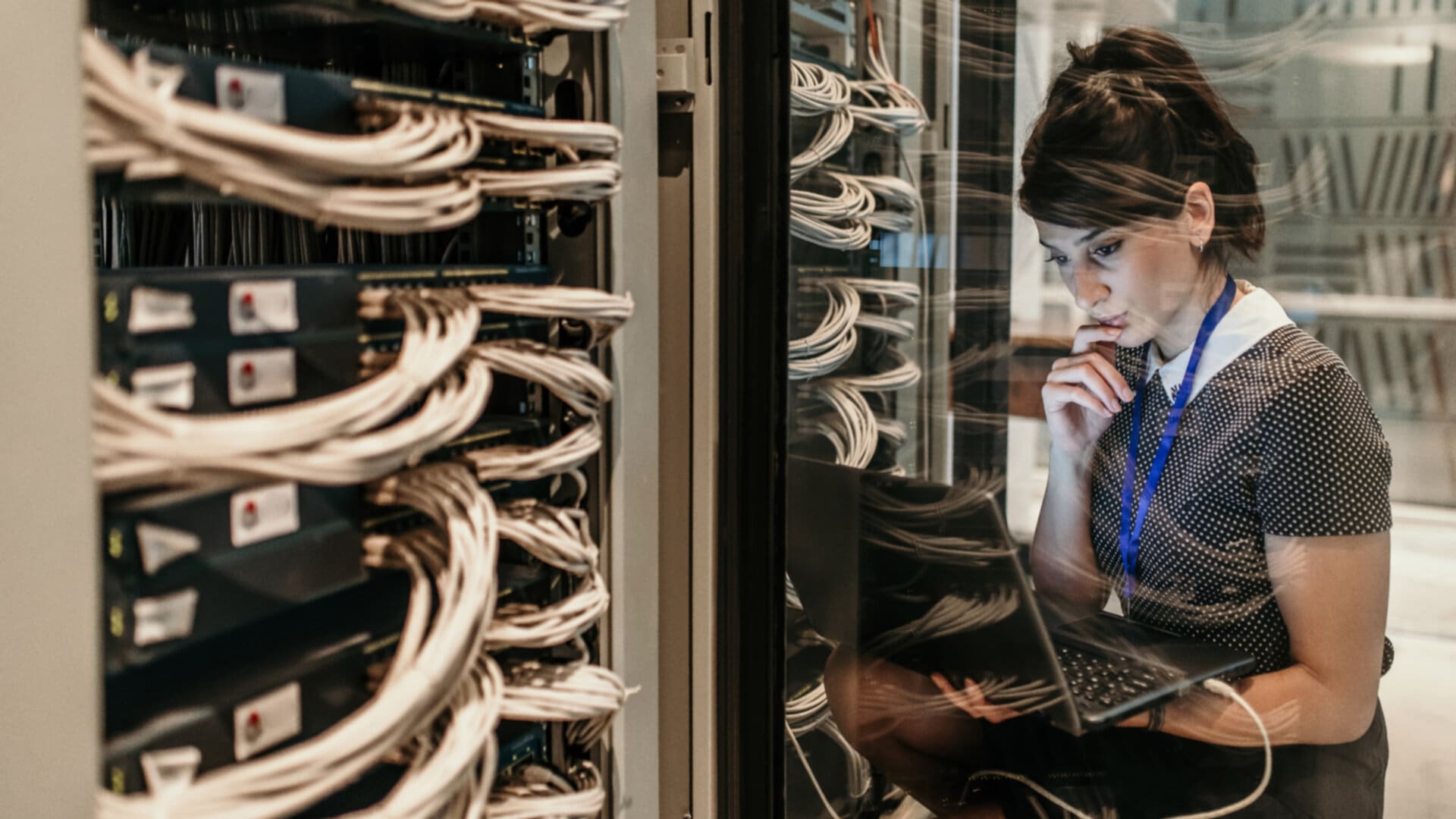 A woman working in a server room. She is looking at a laptop and appears to be troubleshooting a problem.