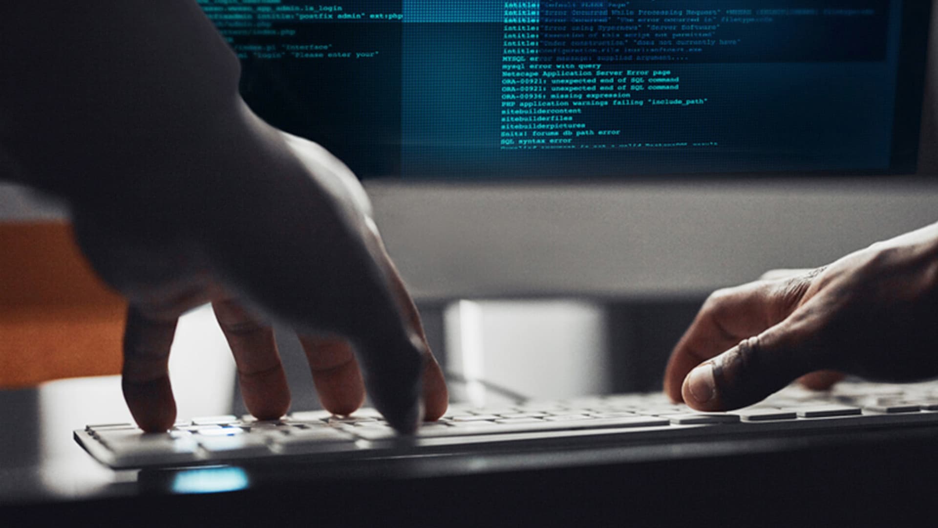 A close-up view of hands typing on a computer keyboard. In the background, a screen is visible displaying lines of code or error messages.