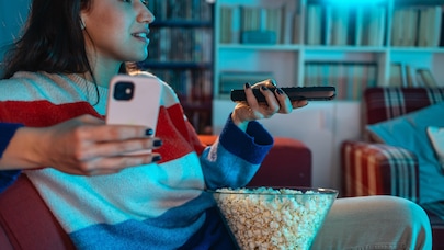 A woman sitting on a couch with a bowl of popcorn in her lap, holding a remote control in her hand while watching TV.