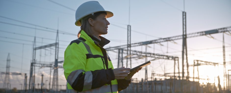 A female engineer wearing a white hard hat and high-visibility jacket stands in an electrical substation, holding a tablet and inspecting the site during sunset.