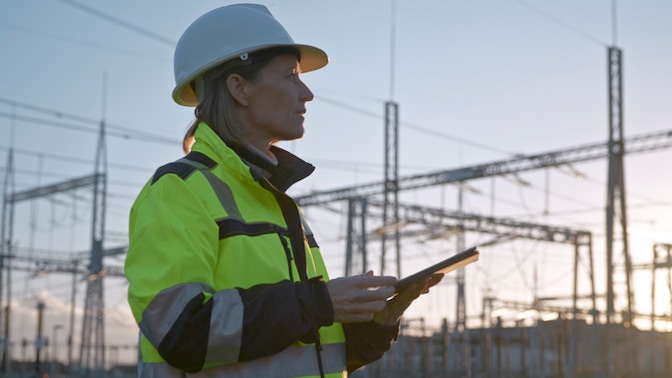 A female engineer wearing a white hard hat and high-visibility jacket stands in an electrical substation, holding a tablet and inspecting the site during sunset.