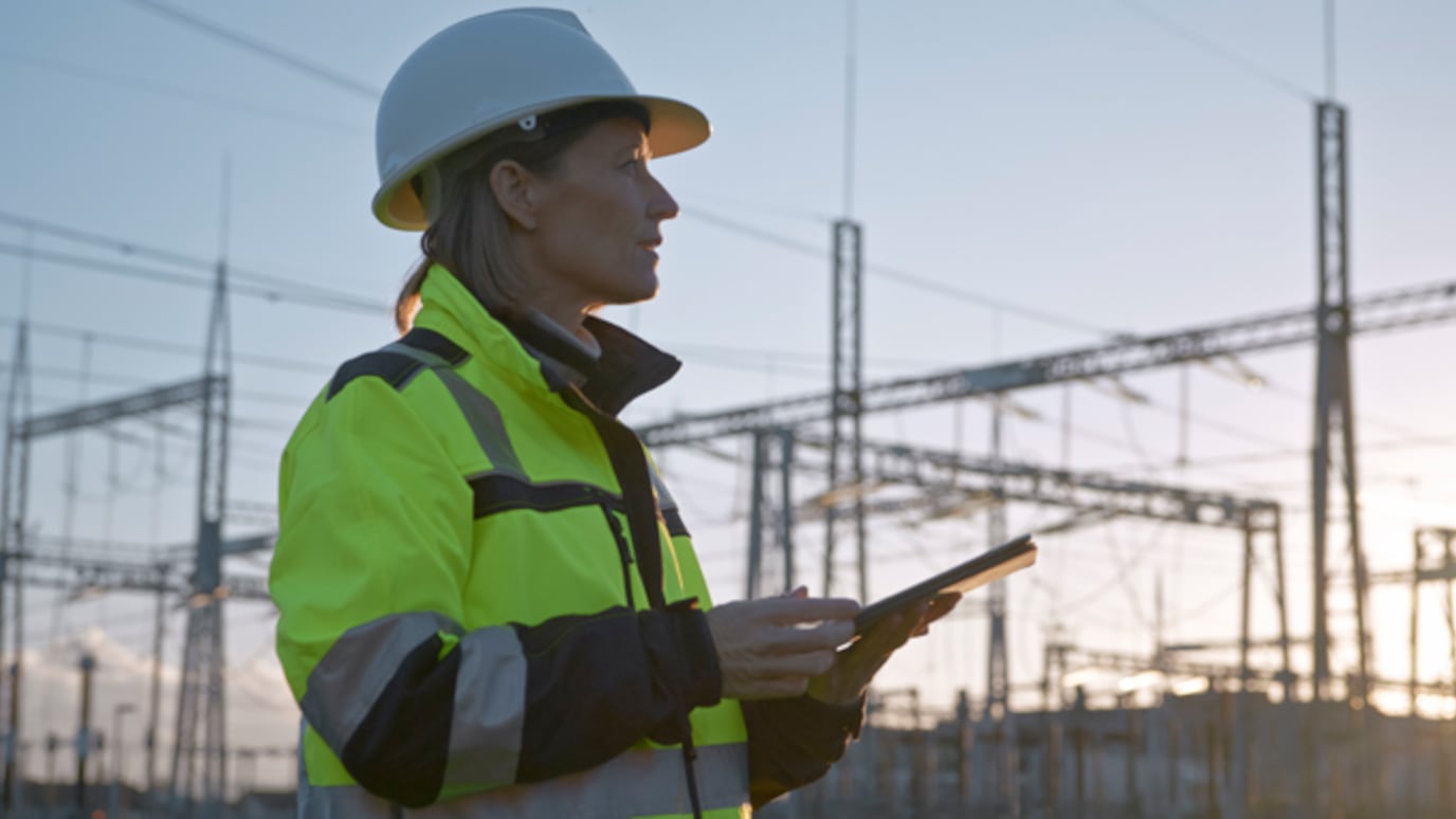 A female engineer wearing a white hard hat and high-visibility jacket stands in an electrical substation, holding a tablet and inspecting the site during sunset.