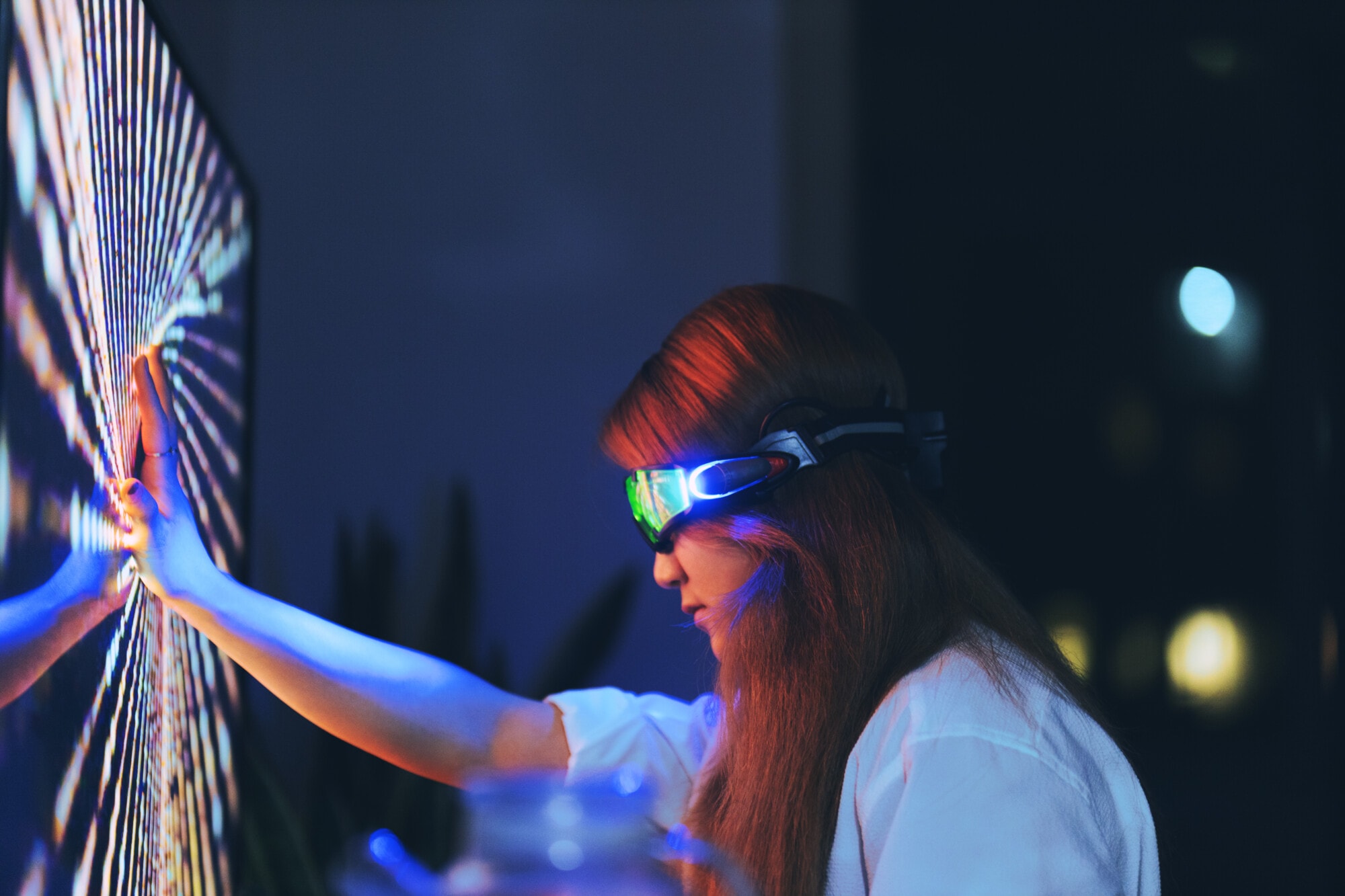 A woman wearing futuristic augmented reality glasses touches a vibrant, glowing digital screen with an outstretched hand in a dark room.