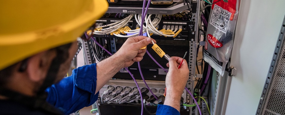 Technician wearing a yellow hard hat and blue uniform labeling cables in a network server rack, ensuring organized cable management.