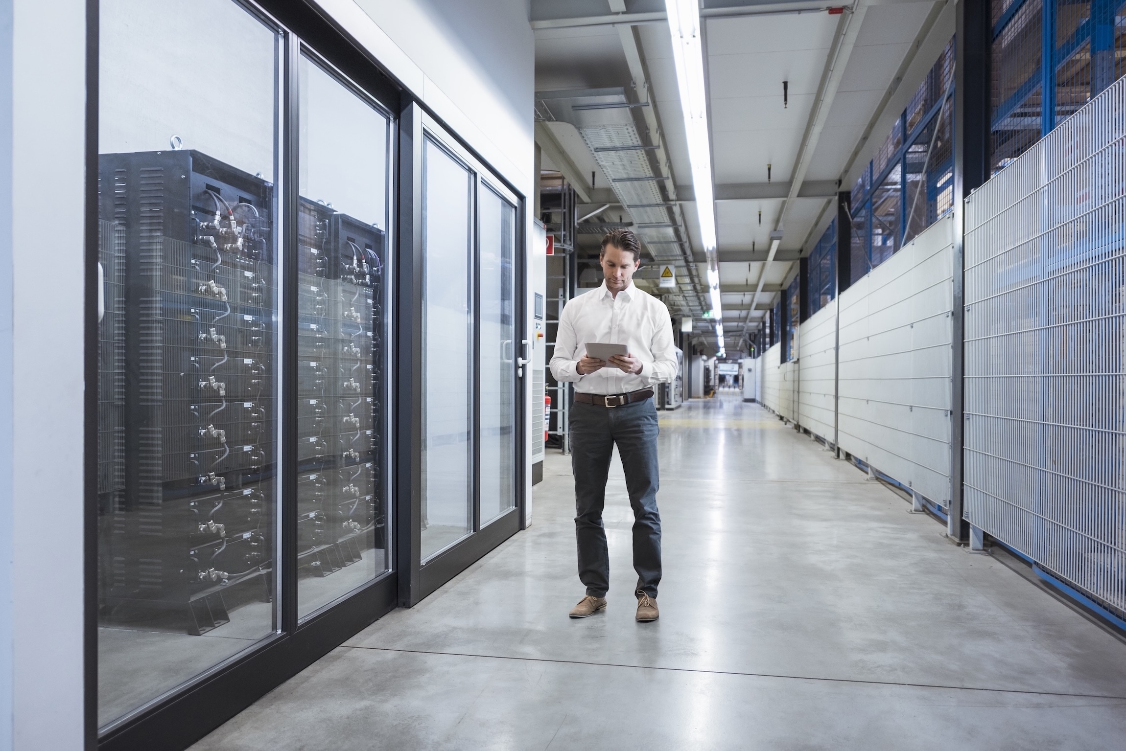 Man in business attire using a tablet while walking through a modern data center corridor with server racks on the side.
