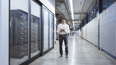 Man in business attire using a tablet while walking through a modern data center corridor with server racks on the side.