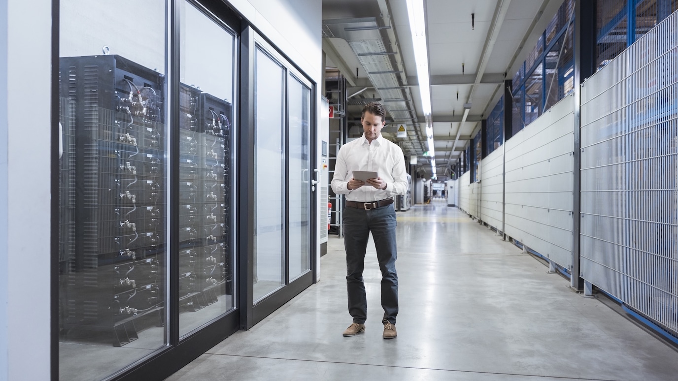 Man in business attire using a tablet while walking through a modern data center corridor with server racks on the side.