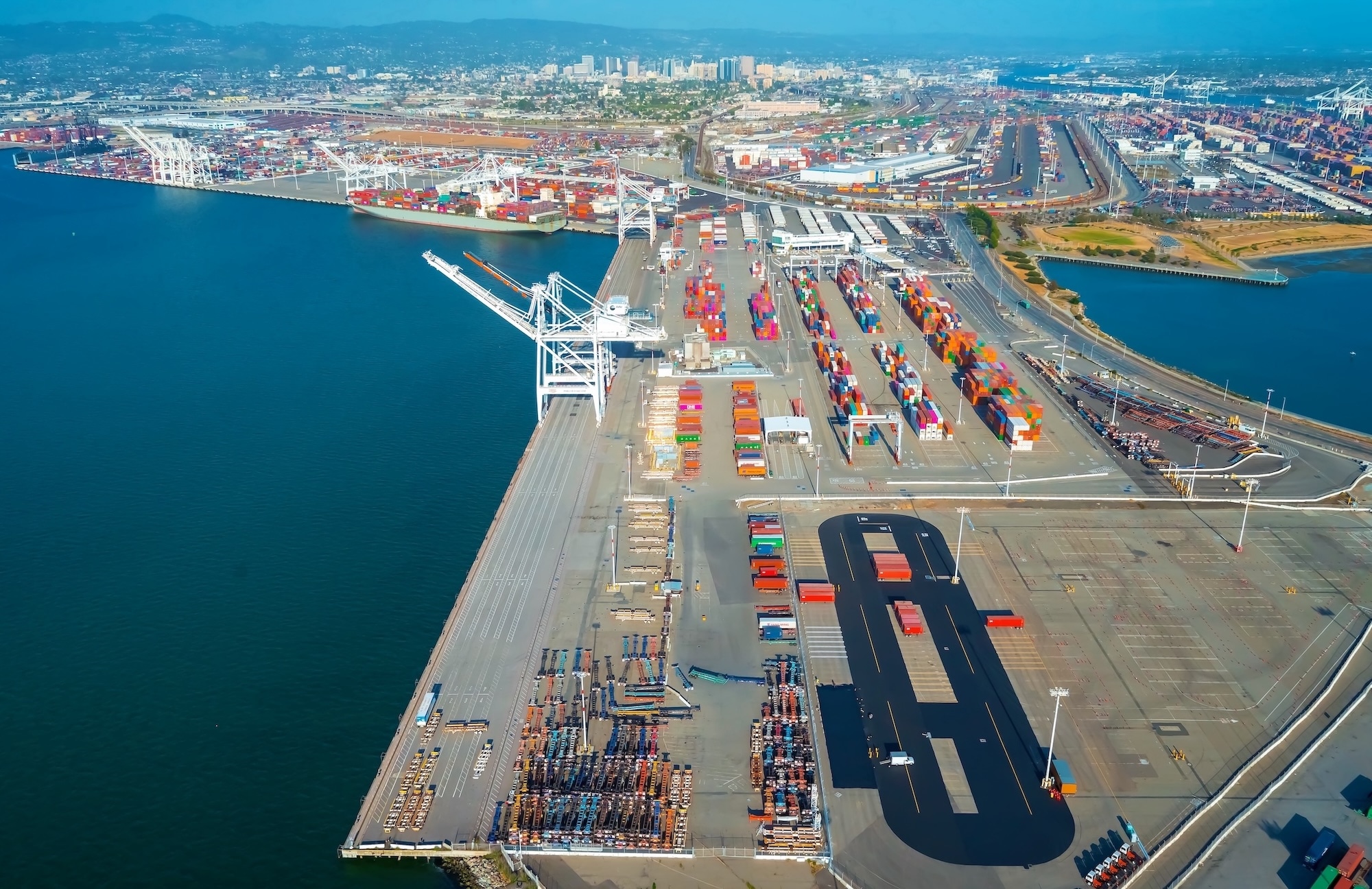 Aerial view of a busy commercial shipping port with cargo containers, cranes, and city skyline in the background.