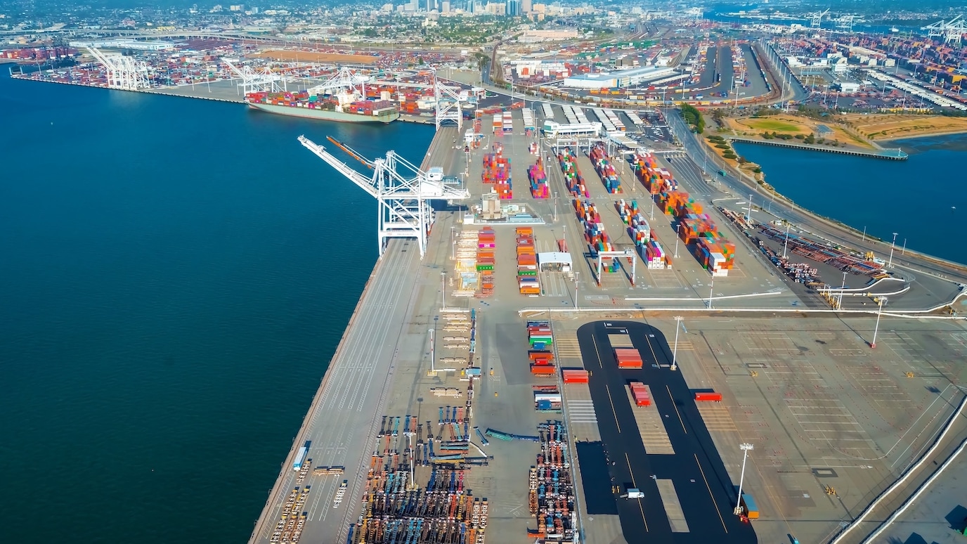 Aerial view of a busy commercial shipping port with cargo containers, cranes, and city skyline in the background.
