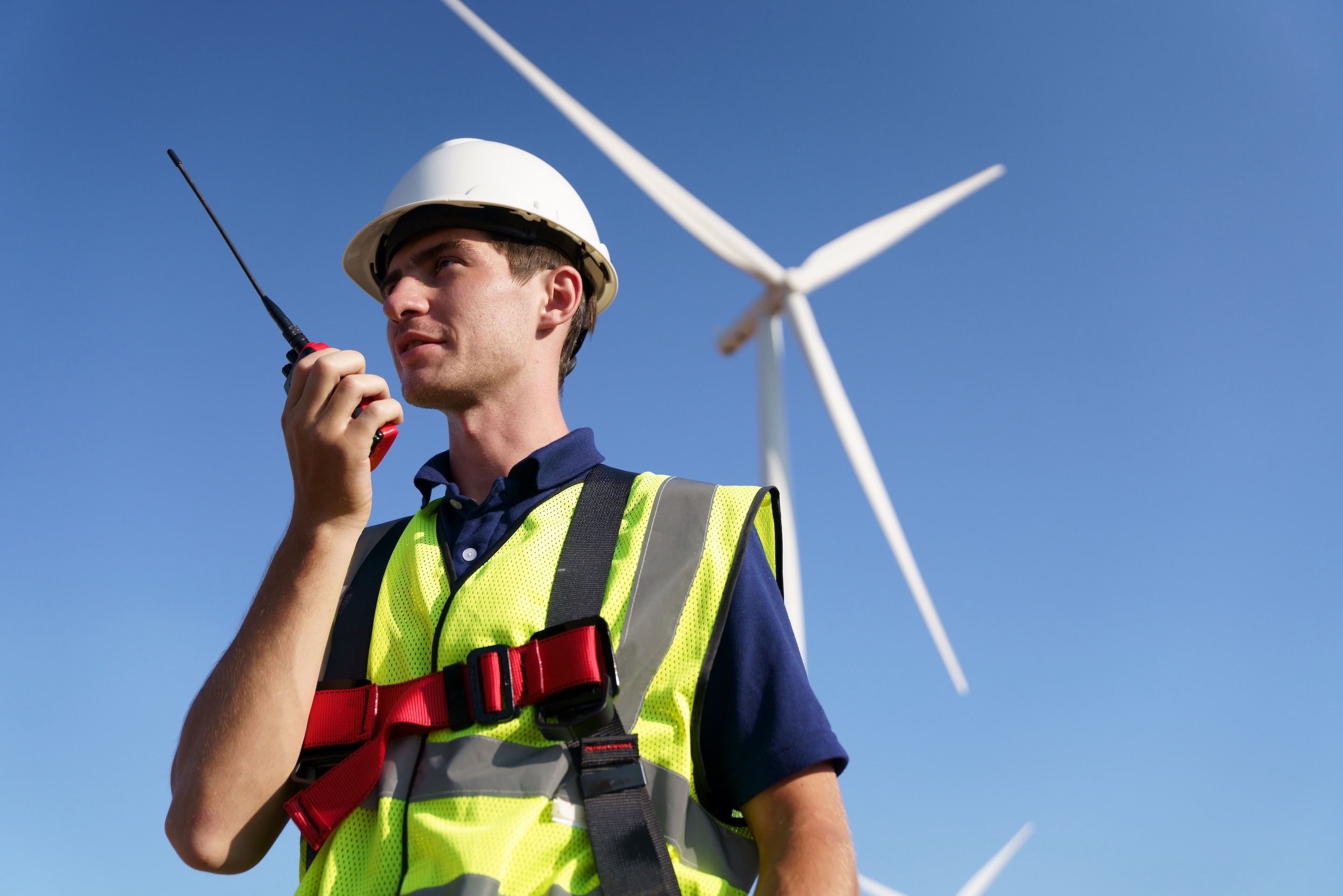Engineer in safety gear uses a walkie-talkie near a wind turbine, overseeing renewable energy site operations.