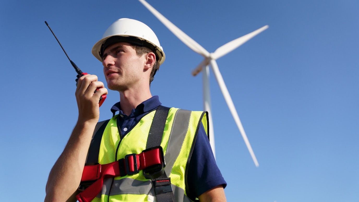 Engineer in safety gear uses a walkie-talkie near a wind turbine, overseeing renewable energy site operations.