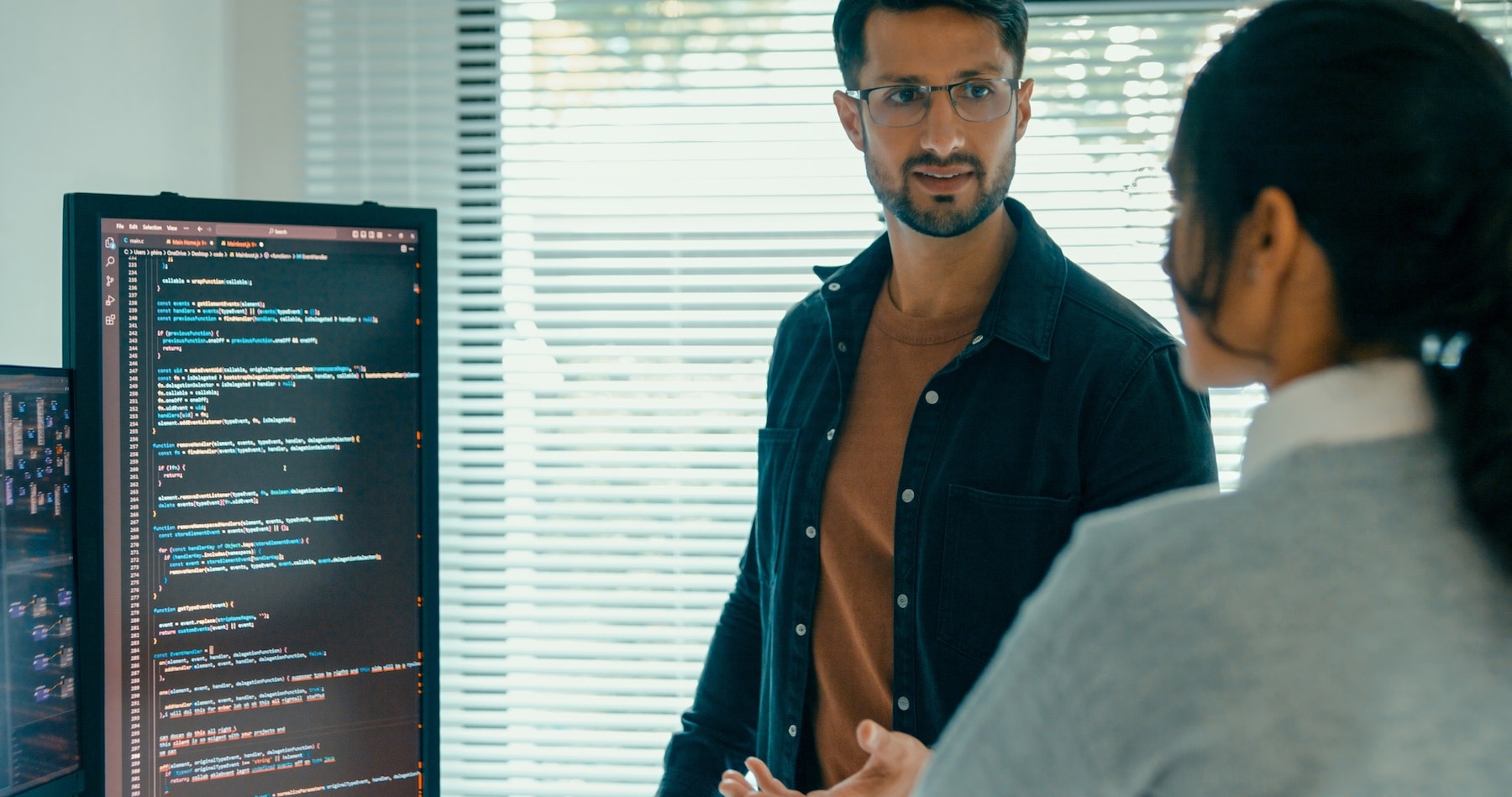 Two software developers discussing code on a screen in a modern office with blinds and natural light in the background.
