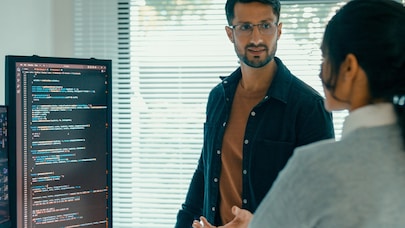Two software developers discussing code on a screen in a modern office with blinds and natural light in the background.