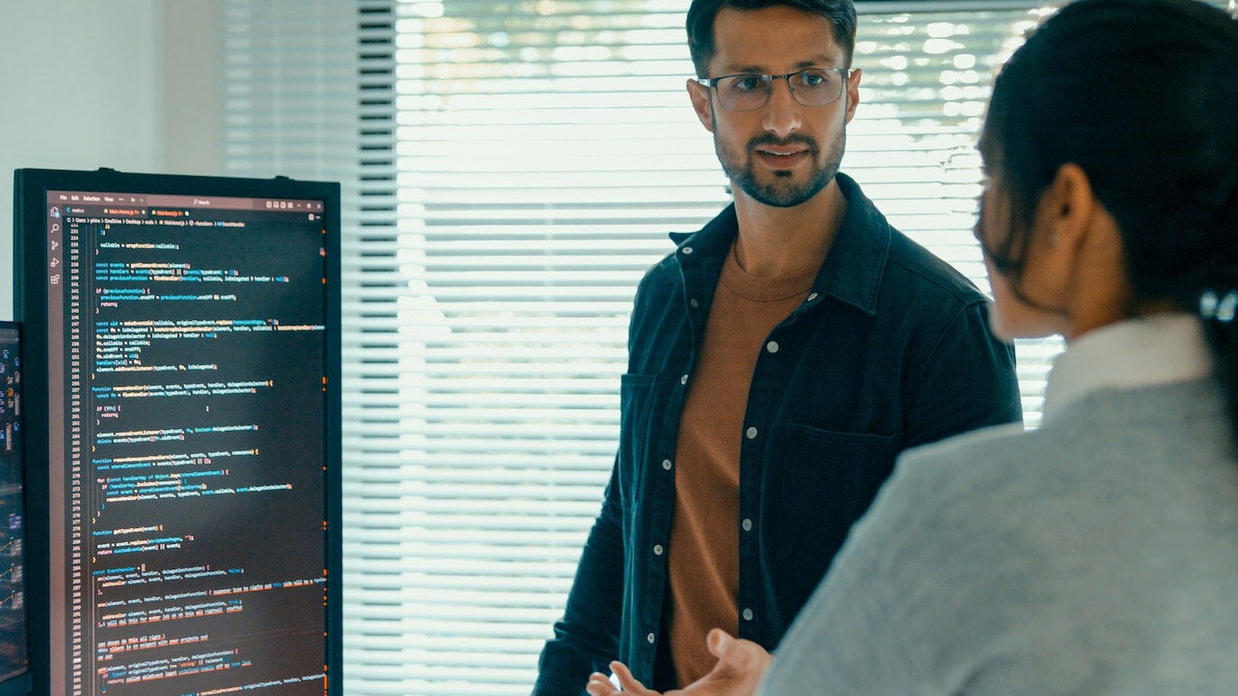 Two software developers discussing code on a screen in a modern office with blinds and natural light in the background.