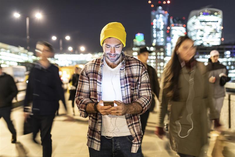 Busy cityscape at night, filled with vibrant lights and a sense of movement. In the foreground, a man is smiling looking at his phone by night with blurred people passing nearby.