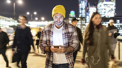 Busy cityscape at night, filled with vibrant lights and a sense of movement. In the foreground, a man is smiling looking at his phone by night with blurred people passing nearby.