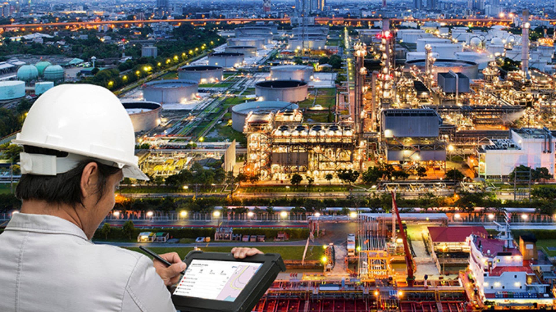 Engineer using a tablet to monitor and manage operations at an illuminated industrial plant with storage tanks and pipelines.