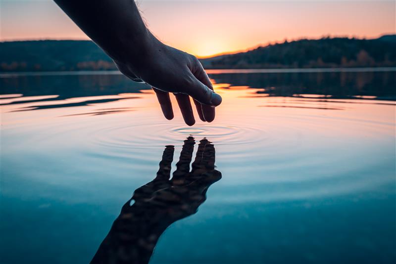 Hand touching calm lake water at sunset, creating ripples with reflection, symbolizing nature, balance, and connection.
