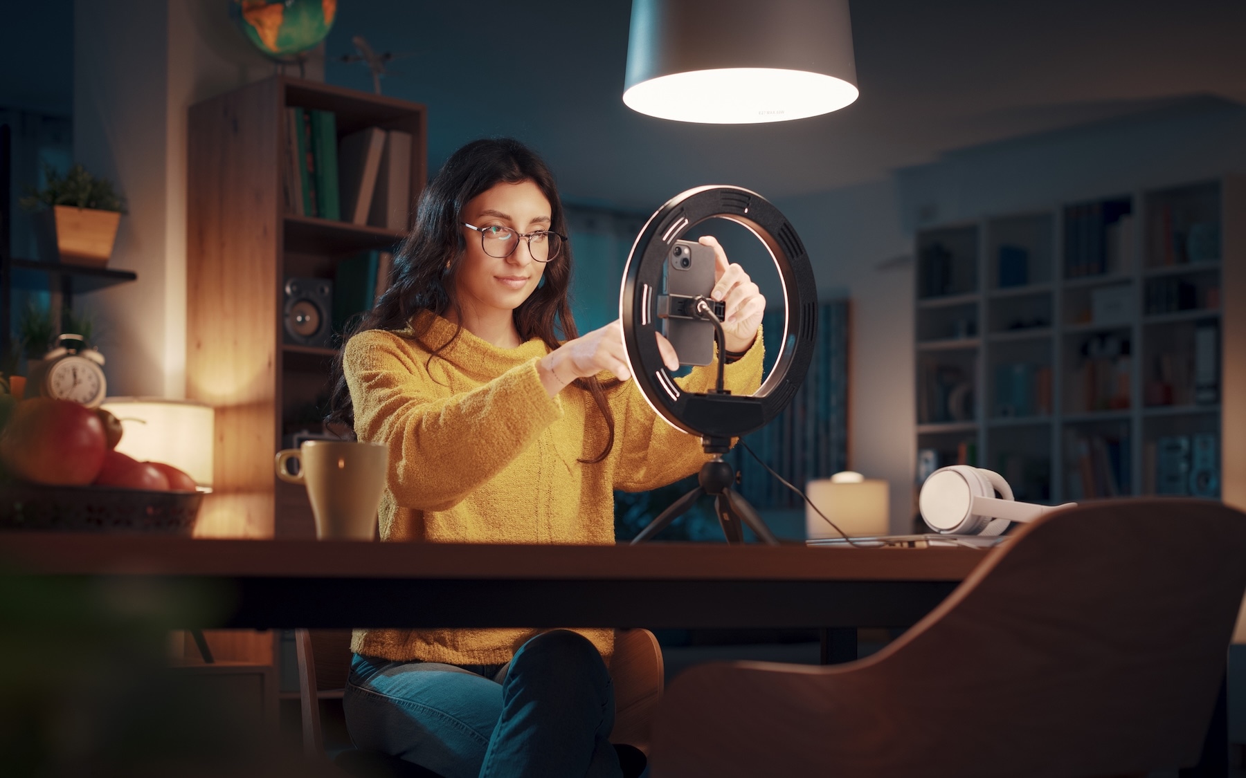 Woman wearing glasses and a yellow sweater adjusts a smartphone on a tripod with a ring light in a cozy room.