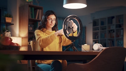 Woman wearing glasses and a yellow sweater adjusts a smartphone on a tripod with a ring light in a cozy room.