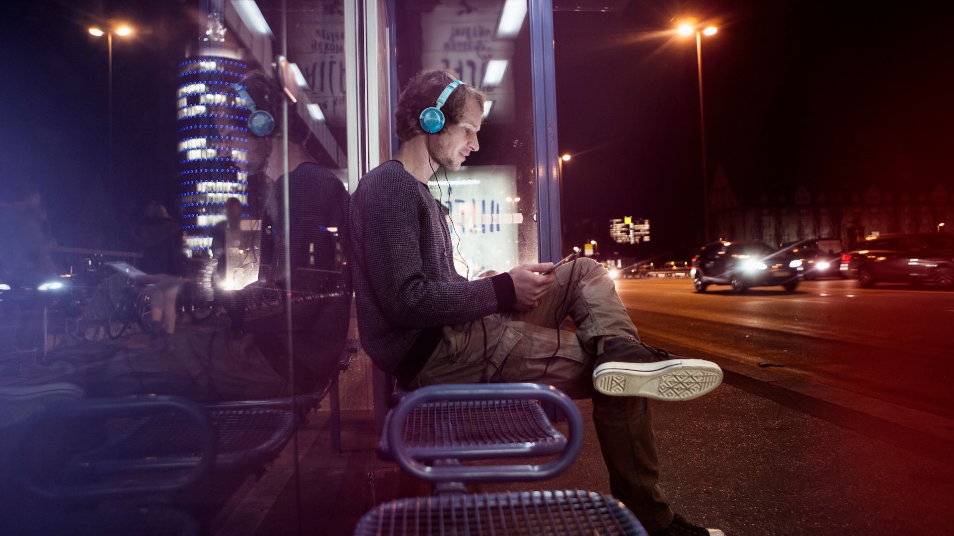 Man sitting at a bus stop wearing headphones and listening to music, enjoying a moment of relaxation in the city.