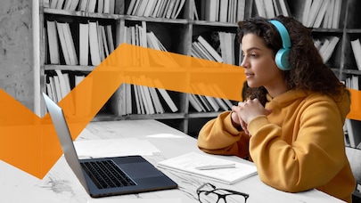 Woman wearing headphones, sat at tabel in library looking at a laptop