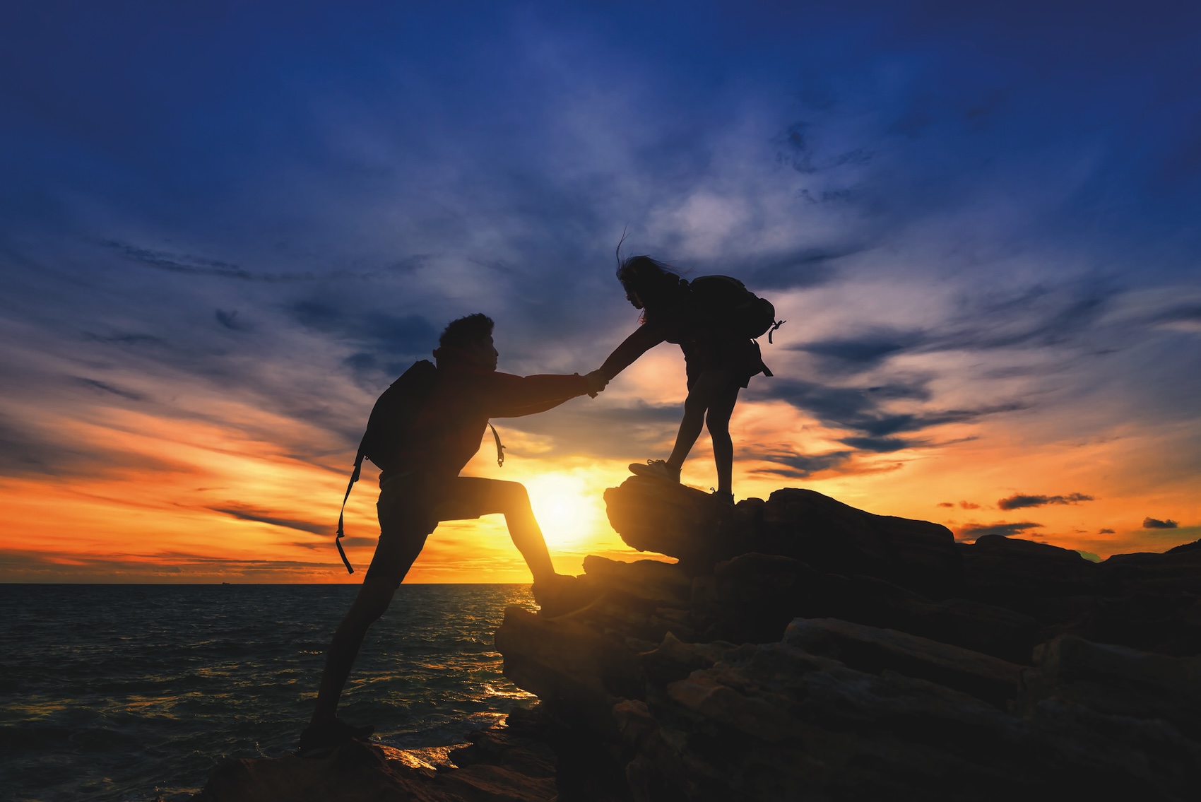 Two hikers silhouetted against a vibrant sunset, one helping the other climb rocks by the sea.