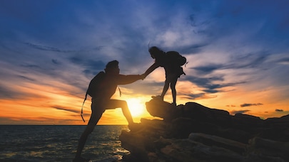 Two hikers silhouetted against a vibrant sunset, one helping the other climb rocks by the sea.