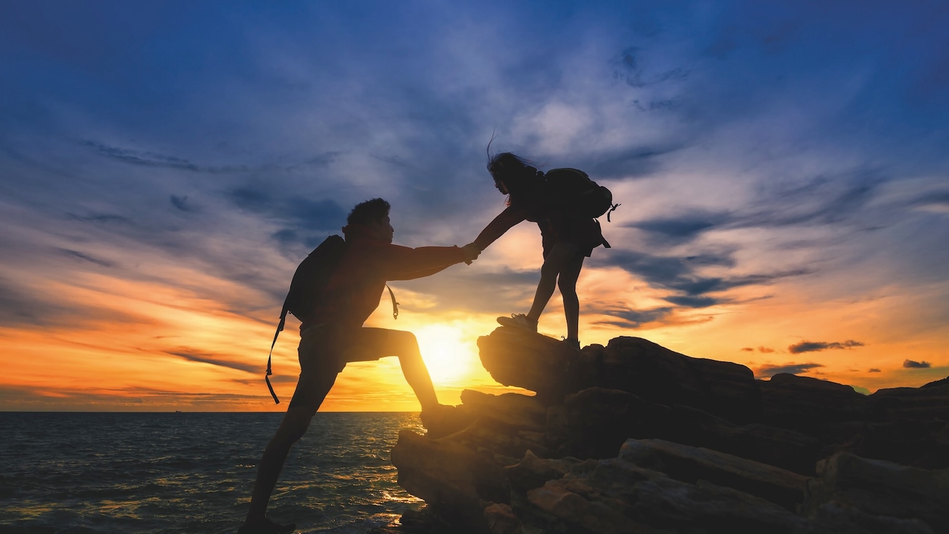 Two hikers silhouetted against a vibrant sunset, one helping the other climb rocks by the sea.