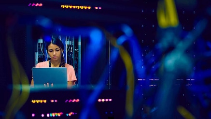 Data center technician looking at laptop, seen through gap in server cables 