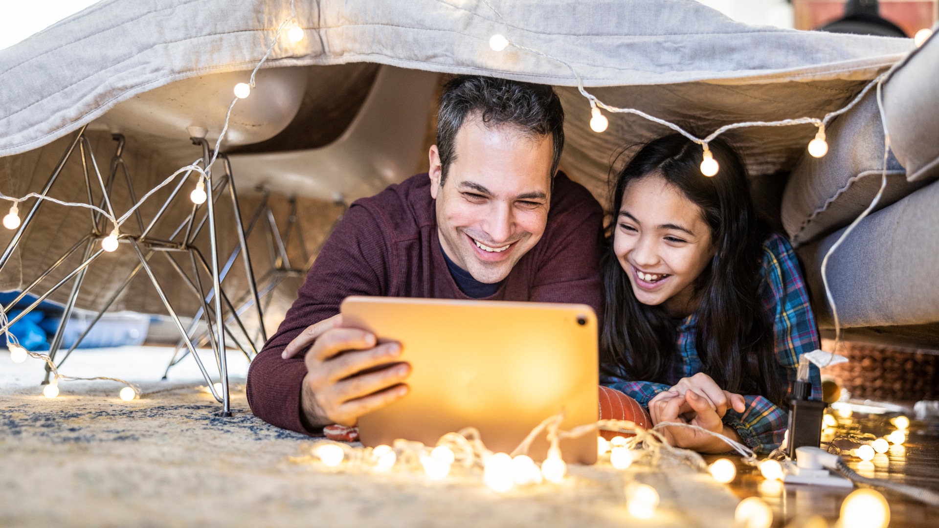 Father and daughter smiling under a blanket fort watching a tablet with string lights at home