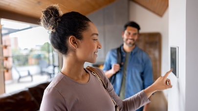Woman adjusting smart thermostat while smiling partner stands in modern home interior