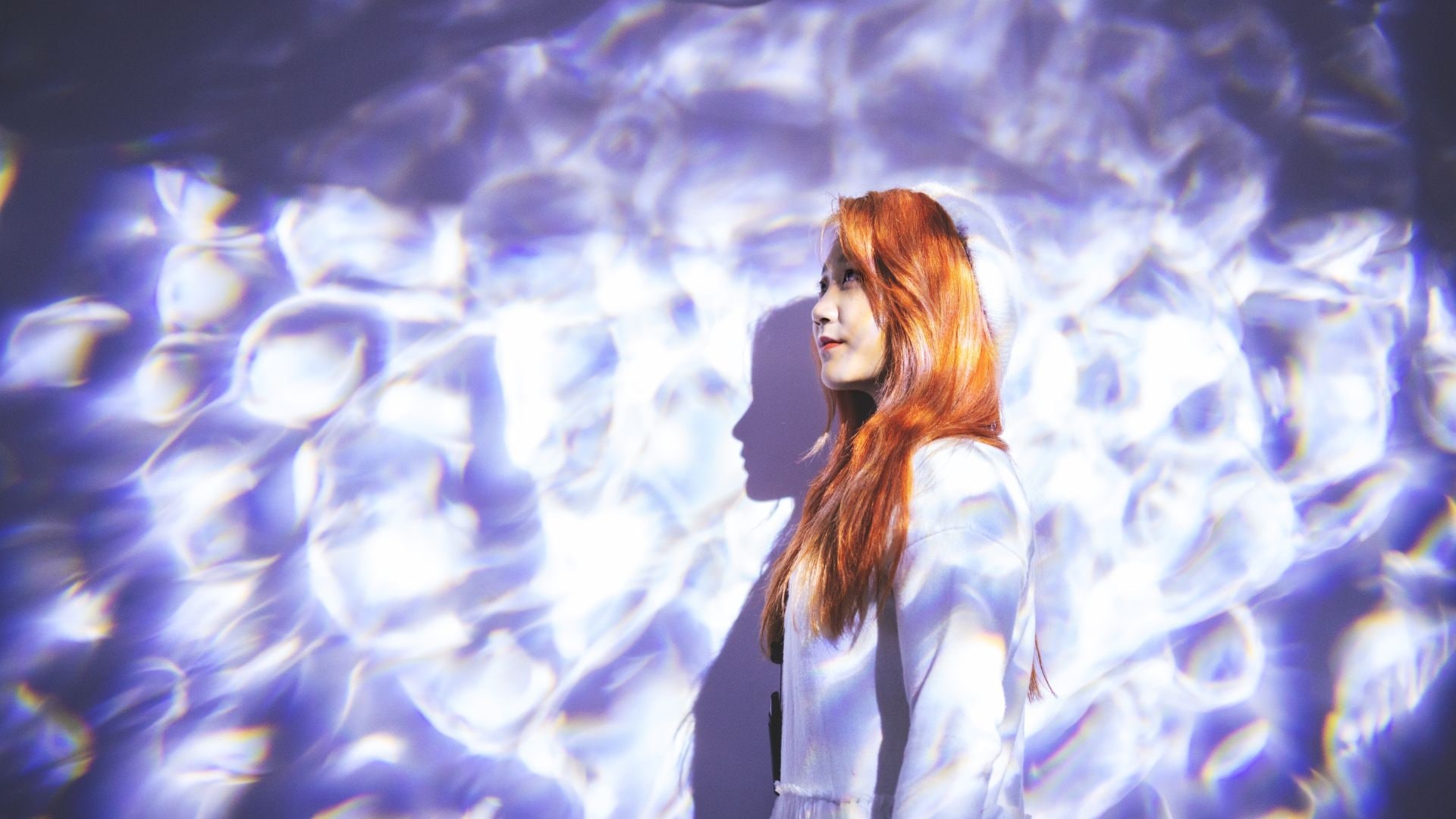 Woman with red hair in profile standing against abstract purple and blue light patterns casting shadows on wall